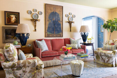 Living room designed by some of The Best Interior Designers and Architects in Birmingham, featuring a red sofa, floral armchairs, blue lamps, wall art, and decorative plates; glass coffee table with books and flowers in front.