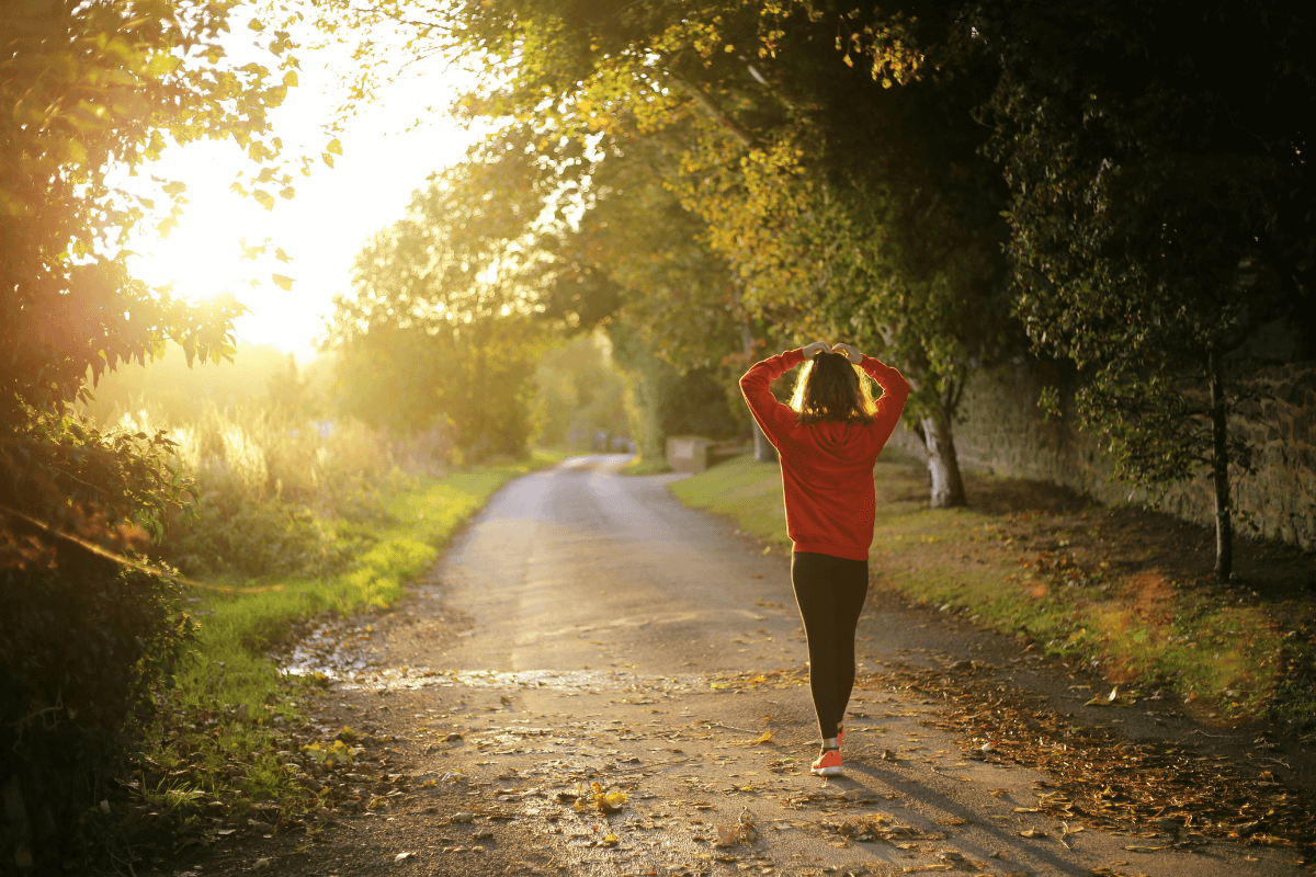 Person in a red sweatshirt walks along a tree-lined path in the sunlight, enjoying the greenery and scattered leaves—a perfect Nashville day for wellness.