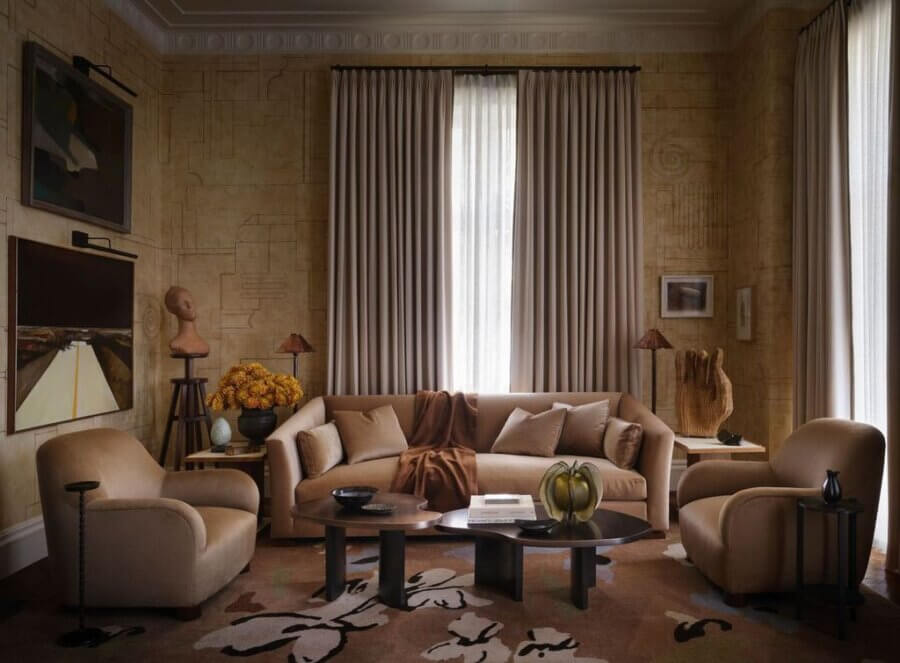 7 Designer Show Houses to Have on Your Radar for 2026 - 6 A warmly lit, modern living room with beige armchairs, a matching sofa, dark coffee tables, floor-length curtains, wall art, and decorative objects on display.