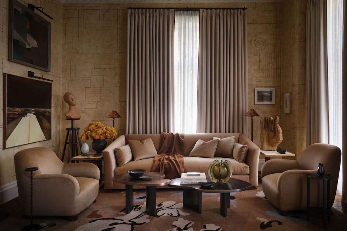 A living room reminiscent of elegant show houses features beige armchairs, a matching sofa with cushions, a dark coffee table, neutral drapes, wall art, and decorative objects in a warm, earthy color palette.