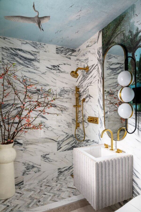 7 Designer Show Houses to Have on Your Radar for 2026 - 3 Modern bathroom with marble walls, gold shower fixtures, a ribbed pedestal sink, a round mirror with globe lights, and ceiling art of a bird and sky.