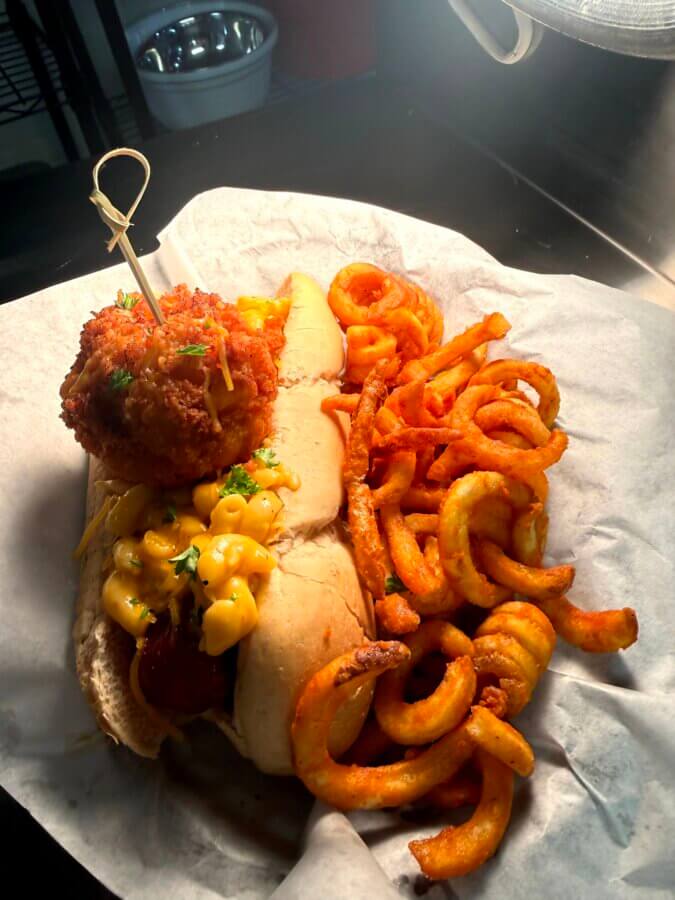 4 New Louisville Restaurants (+2 New Menus!) to Try This Month - 12 A sausage sandwich topped with macaroni and cheese and a fried ball, served with curly fries on parchment paper—a creative dish you’ll find at new Louisville restaurants.
