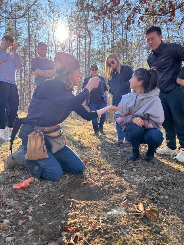 3 Places to Go Truffle Hunting in the South - 3 A group of people outdoors in the South watch as a woman kneels on the ground, showing a truffle to another woman; trees and sunlight are visible in the background.