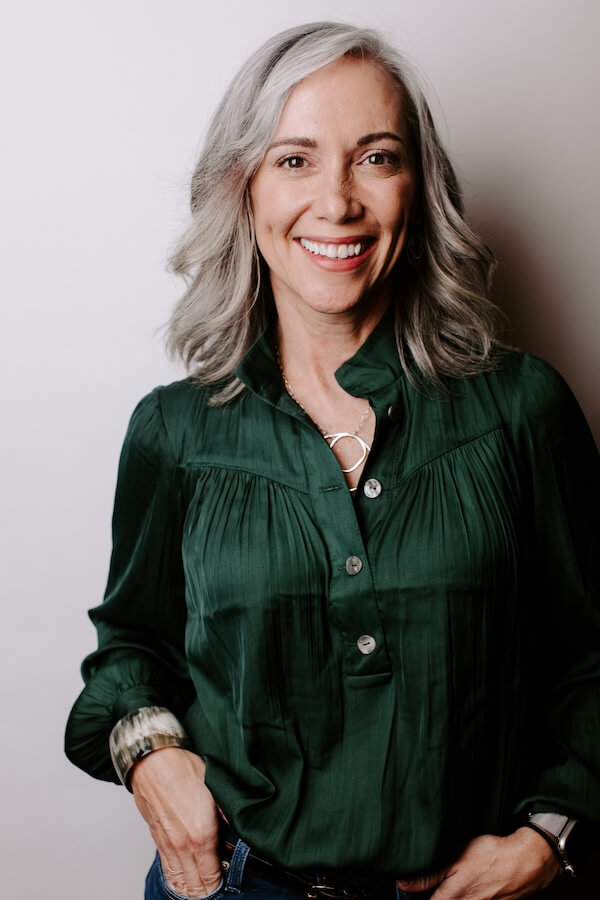 This Nashville Mom Transformed Her Grief Into a Lifeline for Youth - 4 A woman with shoulder-length gray hair, wearing a dark green blouse and blue jeans, stands against a plain light background, smiling with her hands in her pockets—a welcoming presence at the Goldfinch Foundation.