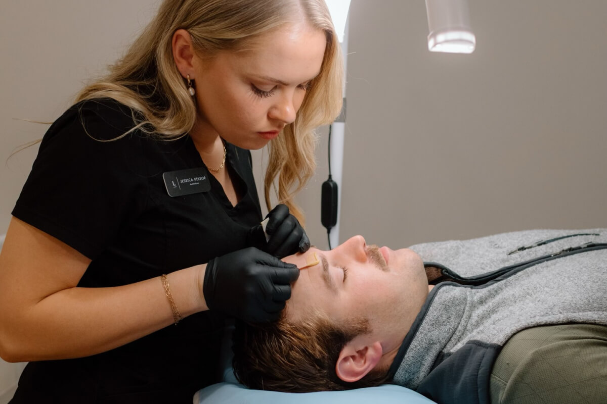 A woman wearing black gloves applies a treatment to a man's forehead as he lies on a medical table—discover how new Nashville services can elevate your everyday skincare routine.