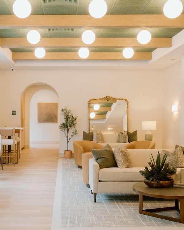 6 New Nashville Services to Elevate Your Everyday - 4 A modern living room with beige sofas, a round coffee table, a large ornate mirror, potted plants, and globe ceiling lights on wooden beams—perfect to elevate everyday relaxation with chic comfort.