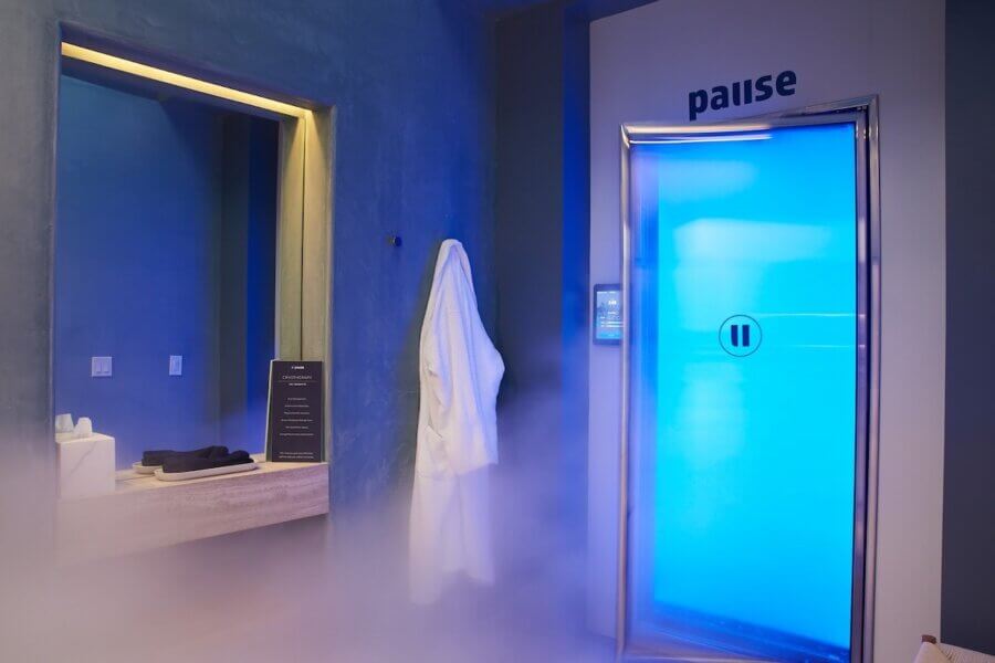 6 New Nashville Services to Elevate Your Everyday - 6 A modern spa room features a blue-lit cryotherapy chamber labeled "pause," a mirror, counter with towels, a sign, and a hanging white robe surrounded by mist—showcasing one of the new Nashville services in relaxation and wellness.