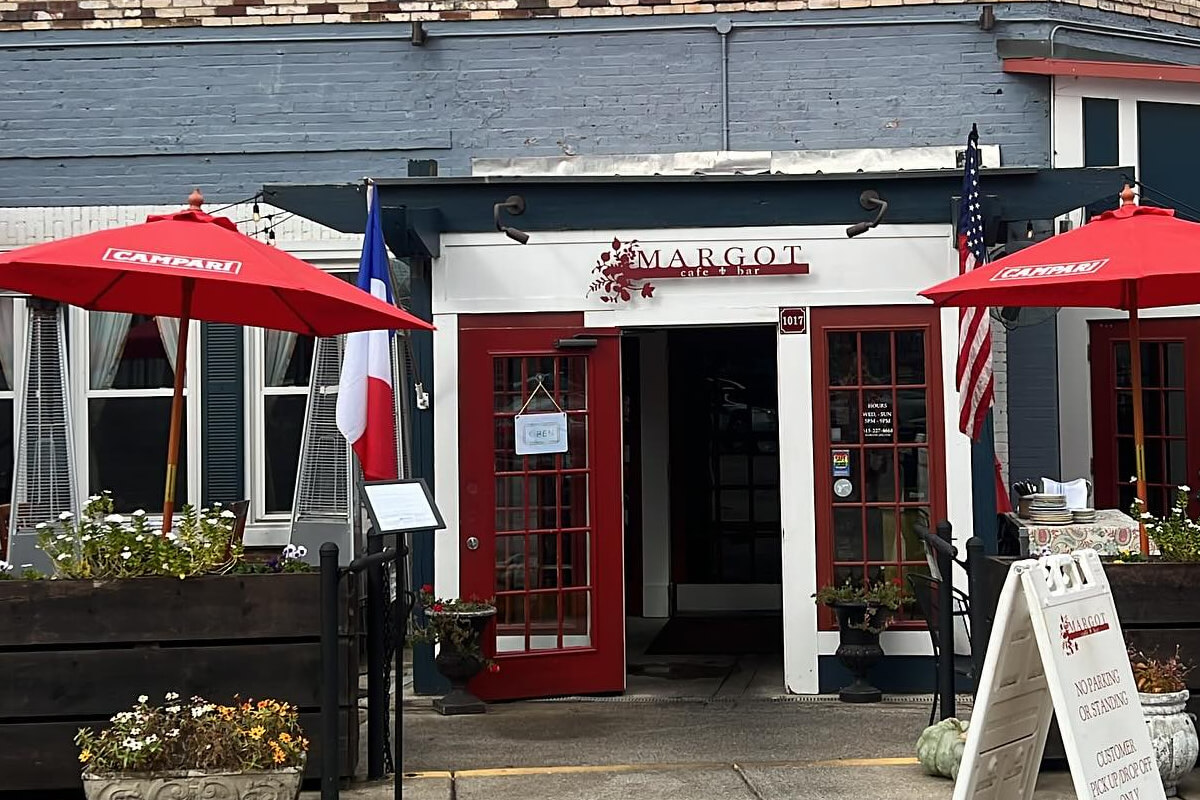 Outdoor entrance to Margot Cafe features red-framed doors, red umbrellas, outdoor seating, and both French and American flags proudly displayed.
