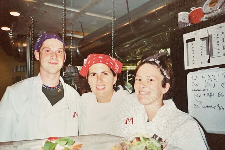 An Ode to Margot Cafe: Au Revoir Mon Amie - 5 Three chefs wearing bandanas and white jackets stand together in the Margot Cafe’s commercial kitchen, smiling at the camera; food dishes are visible in the foreground.