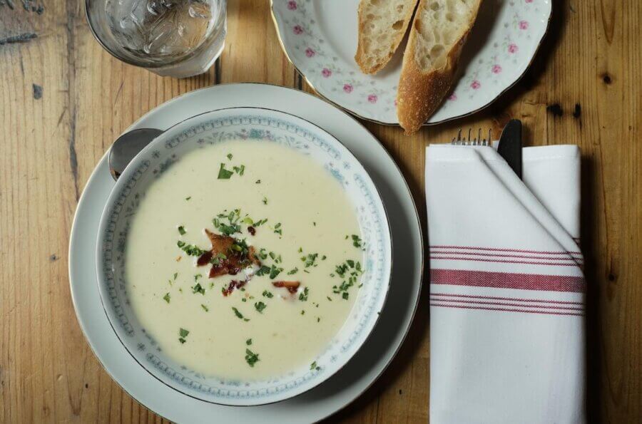 An Ode to Margot Cafe: Au Revoir Mon Amie - 2 A bowl of creamy soup garnished with herbs and bacon sits on a wooden table at Margot Cafe, next to a plate with two bread slices, a glass of water, and a napkin-wrapped fork and knife.