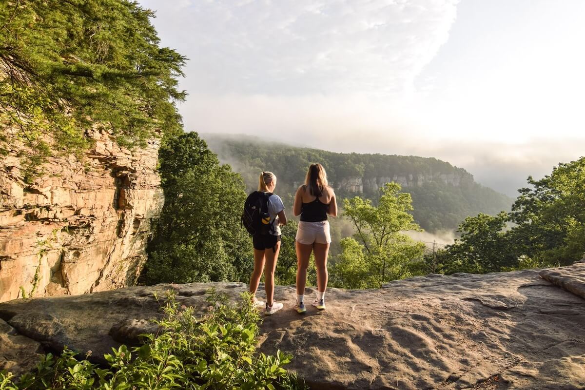 Two adventurous girls stand on a rocky ledge overlooking a forested valley, mist swirling around distant cliffs beneath a partly cloudy sky—perfect for unforgettable Adventurous Girls Trips.