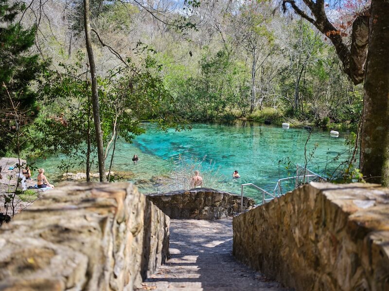 8 Adventurous Girls' Trips (All Skill Levels!) - 5 Stone steps lead down to a clear natural spring where several people are swimming and relaxing; trees and greenery surround the water—an ideal spot for adventurous girls trips seeking fun and relaxation in nature.