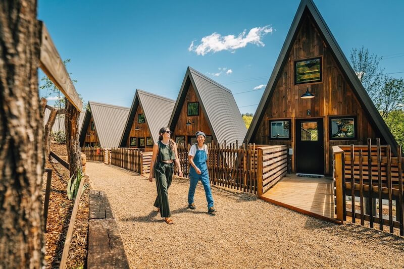 8 Adventurous Girls' Trips (All Skill Levels!) - 10 Two people walk on a gravel path in front of modern A-frame cabins with wood and metal exteriors—an ideal spot for adventurous girls trips on a sunny day.