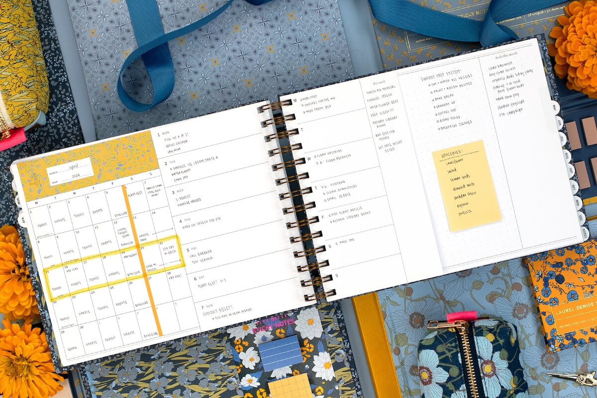 An open Paper Planner Pro with monthly and weekly layouts is surrounded by floral-patterned notebooks, fabric swatches, and yellow flowers on a blue background.