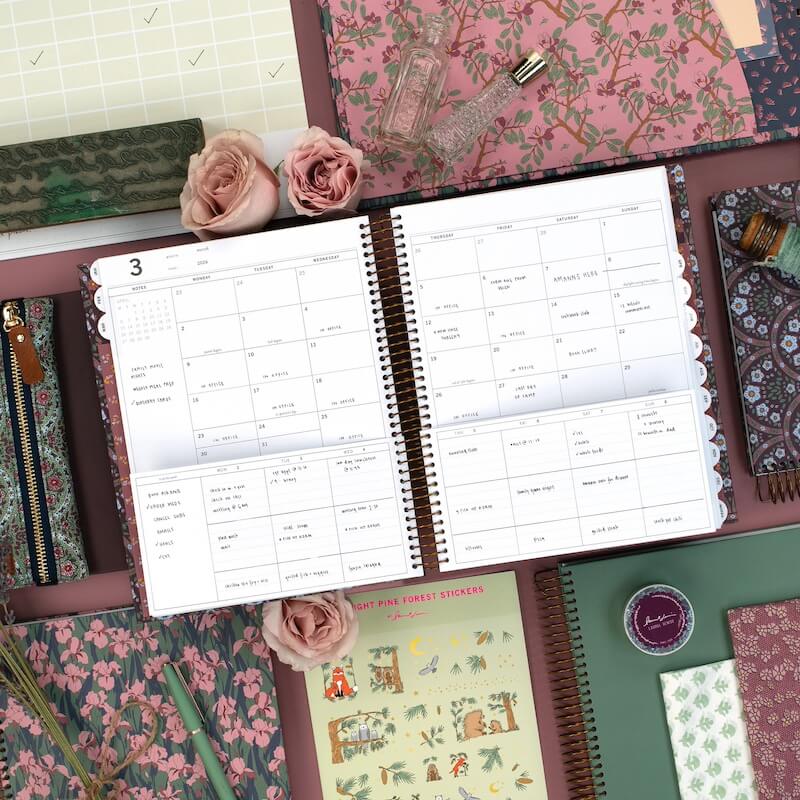 The 2026 Life Hack You Need? A Laurel Denise Planner - 3 A Laurel Denise Planner open to a monthly calendar spread is surrounded by floral-patterned stationery, notebooks, pens, roses, and decorative items on a pink surface—perfect for your 2026 life hack organization.