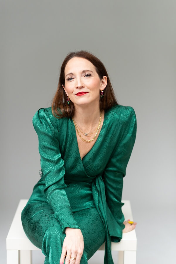 The 2026 Life Hack You Need? A Laurel Denise Planner - 2 Woman in a shiny green wrap dress sits on a white stool against a plain gray background, looking at the camera with a slight smile.