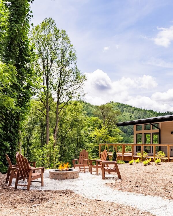 8 Adventurous Girls' Trips (All Skill Levels!) - 7 Four wooden chairs surround a lit fire pit in a landscaped outdoor area beside a modern building—an inviting spot for adventurous girls trips, with trees and hills in the background under a partly cloudy sky.