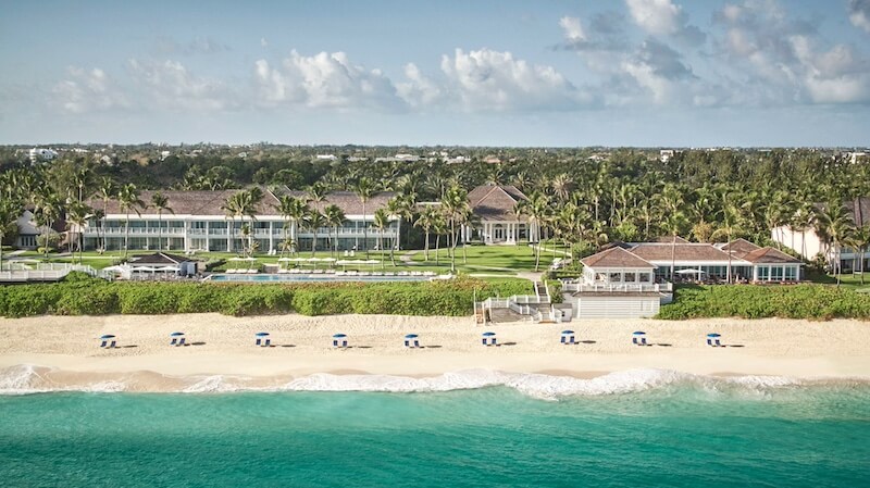 The Unexpected Bahamas Trip You Need To Take - 4 Aerial view of a beachfront resort in the Bahamas with white buildings, palm trees, green lawns, and blue umbrellas on the sandy shore by turquoise water—perfect for your dream Bahamas vacation.