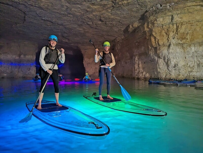 8 Adventurous Girls' Trips (All Skill Levels!) - 2 Two people stand on paddleboards with paddles in hand, illuminated by colored lights, inside a large underground cave with rocky walls—perfect for adventurous girls trips seeking unique and thrilling experiences.