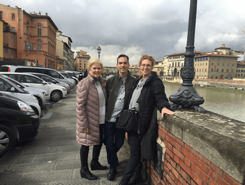 Meet Staci Davidman of Atlanta's Beloved Foxgloves Antiques - 3 Three people stand and pose by a stone railing along a river in a European city, with parked cars and historic buildings under a cloudy sky.