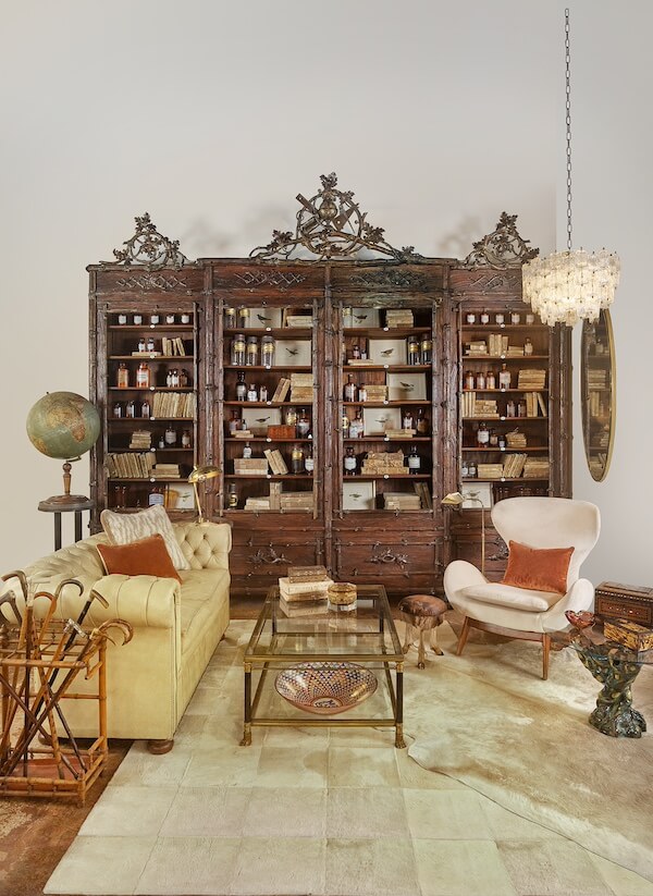 Meet Staci Davidman of Atlanta's Beloved Foxgloves Antiques - 5 A vintage room with a large ornate wooden bookshelf filled with bottles, a beige sofa, armchair, glass coffee table, globe, chandelier, and decorative items from foxglove antiques.