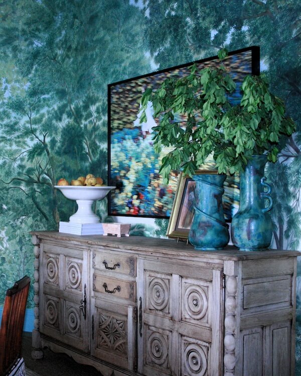 Meet Staci Davidman of Atlanta's Beloved Foxgloves Antiques - 8 A rustic wooden sideboard from foxglove antiques holds a white fruit bowl, blue vases with green branches, and framed art; a TV above displays a pixelated nature scene, with tree wallpaper in the background.