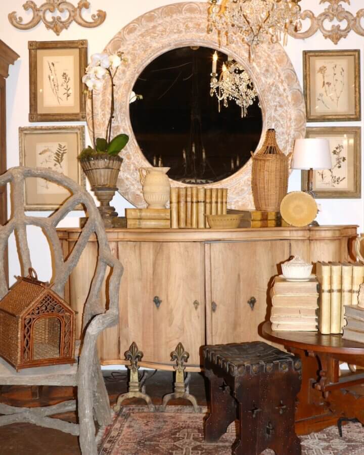 Meet Staci Davidman of Atlanta's Beloved Foxgloves Antiques - 6 A rustic room with a wooden sideboard, round mirror, framed botanical prints from Foxglove Antiques, vintage books, wicker decor, and a carved wooden chair.
