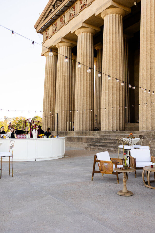 A Gold-Washed Wedding at Nashville's Iconic Parthenon - 4 Outdoor event setup with lounge chairs, tables, and a bar in front of a large building with tall columns and string lights overhead, perfect for a Golden Parthenon wedding.