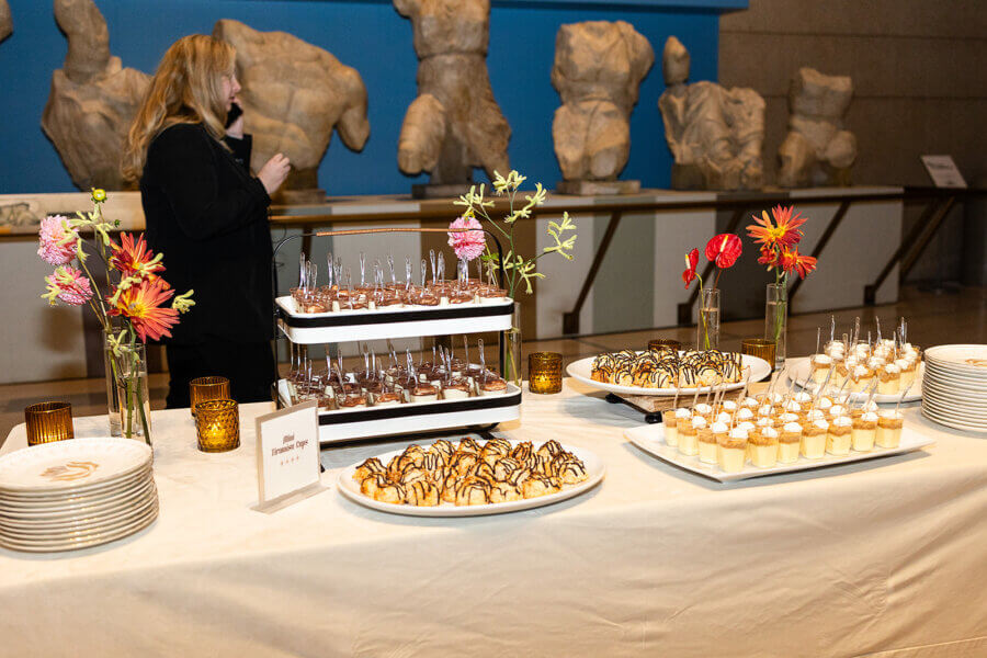 A Gold-Washed Wedding at Nashville's Iconic Parthenon - 14 A table with assorted desserts, including pastries and parfaits, is set up for a Golden Parthenon wedding; a woman stands behind the table near classical statues.
