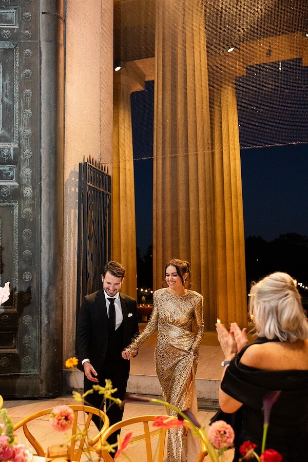 A Gold-Washed Wedding at Nashville's Iconic Parthenon - 13 A couple dressed formally, the woman in a gold gown and the man in a suit, walk hand-in-hand into an elegant Parthenon wedding venue as guests applaud. Tall columns and warm lighting are visible.