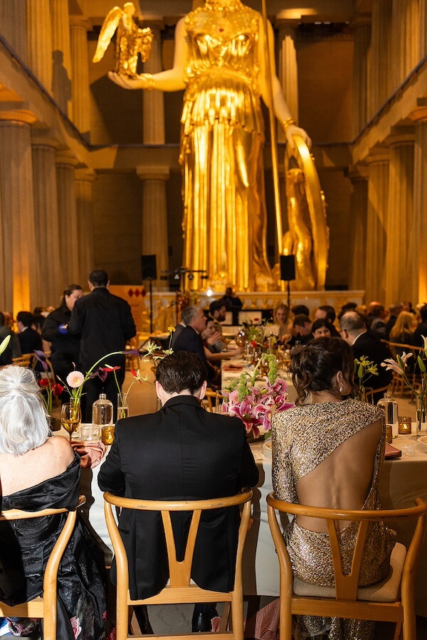 A Gold-Washed Wedding at Nashville's Iconic Parthenon - 15 Guests sit at round tables during a formal dinner event, surrounded by tall columns, in front of a large golden Athena statue—perfect for a Golden Parthenon wedding in this grand hall.