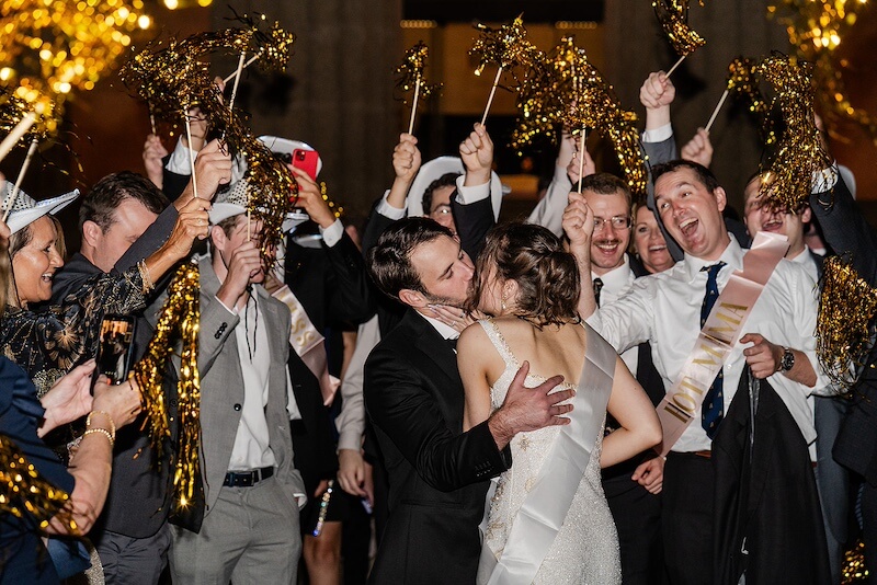 A Gold-Washed Wedding at Nashville's Iconic Parthenon - 19 A bride and groom share a kiss as guests cheer and wave gold pom-poms, creating a vibrant Golden Parthenon wedding celebration.