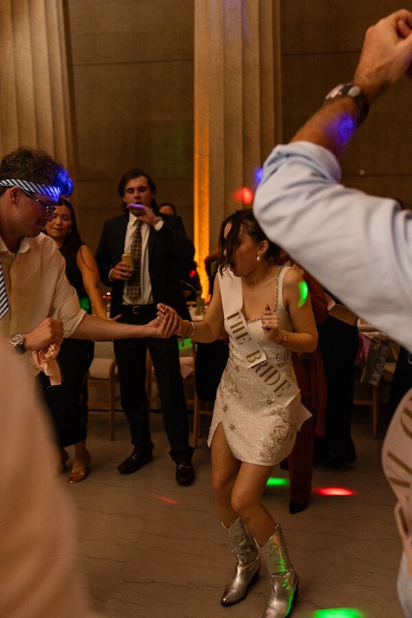 A Gold-Washed Wedding at Nashville's Iconic Parthenon - 18 A woman wearing a "THE BRIDE" sash and silver boots dances with a man at a Golden Parthenon wedding, surrounded by other guests in formal attire.