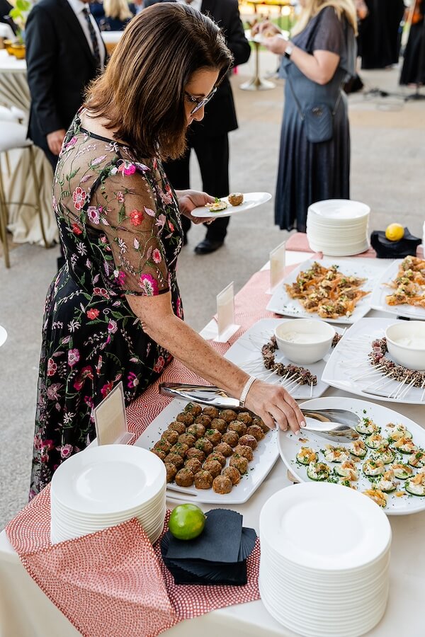 A Gold-Washed Wedding at Nashville's Iconic Parthenon - 11 At a Golden Parthenon wedding, a woman in a floral dress serves herself food from a buffet table with plates of appetizers, bowls, and neatly stacked dishes.