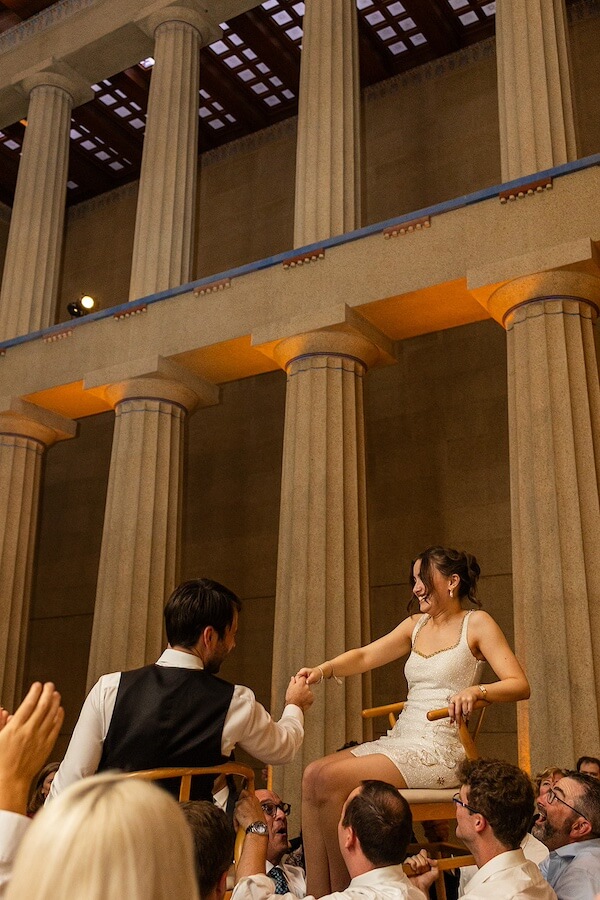 A Gold-Washed Wedding at Nashville's Iconic Parthenon - 16 A couple sits on chairs held aloft during a parthenon wedding celebration in a grand hall with tall columns, holding hands and smiling at each other.