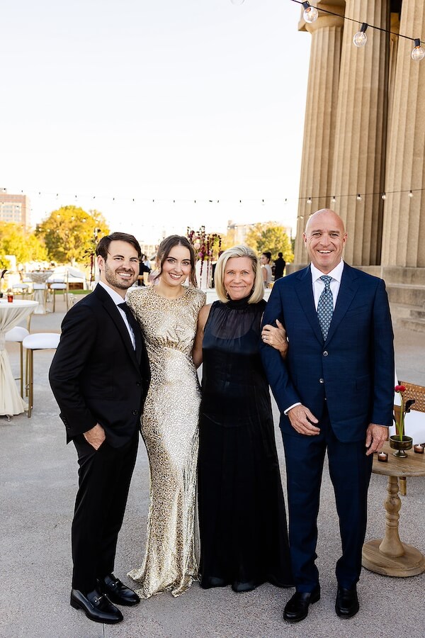 A Gold-Washed Wedding at Nashville's Iconic Parthenon - 3 Four people dressed in formal attire stand together outdoors, smiling at the camera during a Parthenon wedding, with tables and string lights visible in the background.