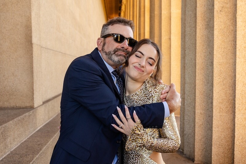 A Gold-Washed Wedding at Nashville's Iconic Parthenon - 8 A man in a suit and sunglasses hugs a woman in a gold outfit. They stand close together by a stone columned wall, both appearing content at their Parthenon wedding.