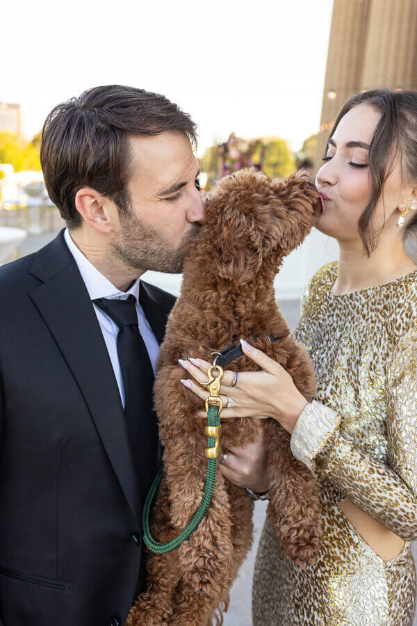 A Gold-Washed Wedding at Nashville's Iconic Parthenon - 12 A man and woman, dressed formally for their Golden Parthenon wedding, kiss a brown dog on either side of its face while holding its leash outdoors.