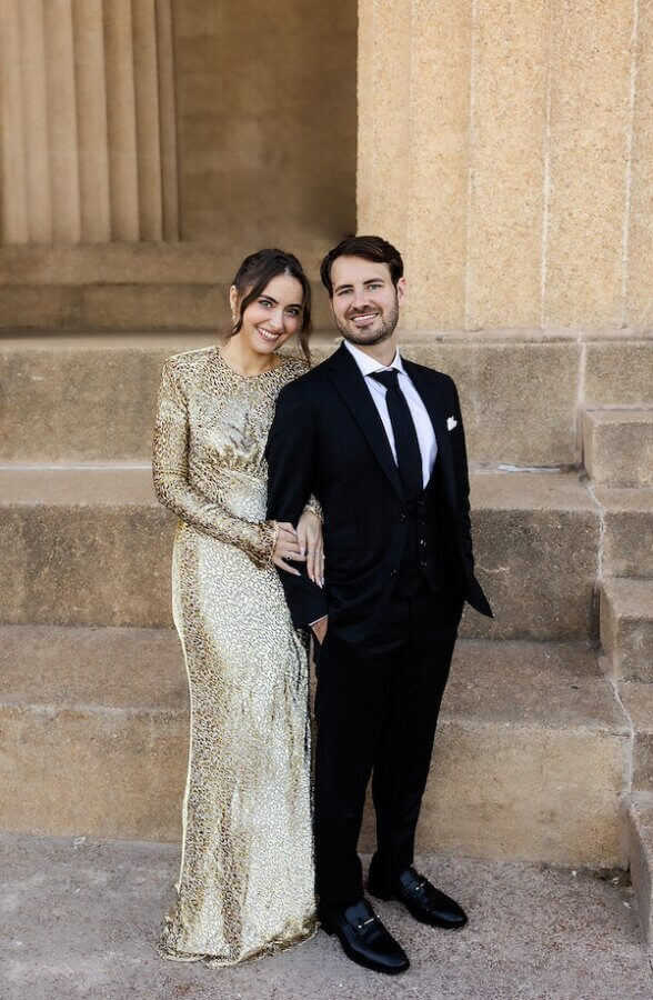 A Gold-Washed Wedding at Nashville's Iconic Parthenon - 2 Woman in a gold sequin dress stands next to a man in a black suit and tie; both are smiling and posing on stone steps, capturing their Golden Parthenon wedding in front of an elegant columned building.