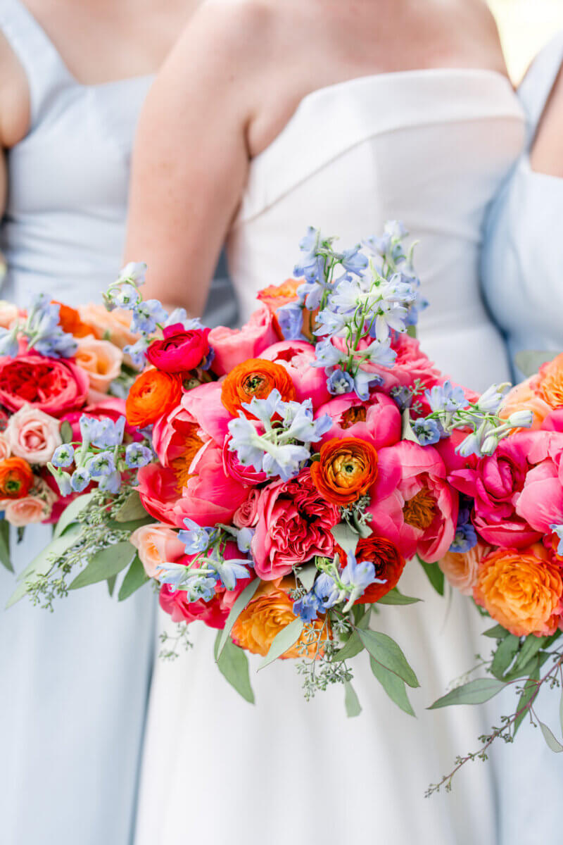 Three people in pastel dresses hold bright bouquets from Birmingham florists, bursting with pink, red, orange, and blue flowers. Only the upper bodies and flowers are visible.