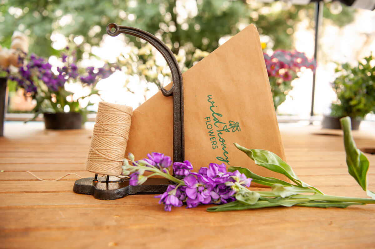 A Wild Yarrow Flowers envelope, a spool of twine, and purple flowers rest on a wooden table with plants in the background, evoking the charming artistry found at Birmingham florists.