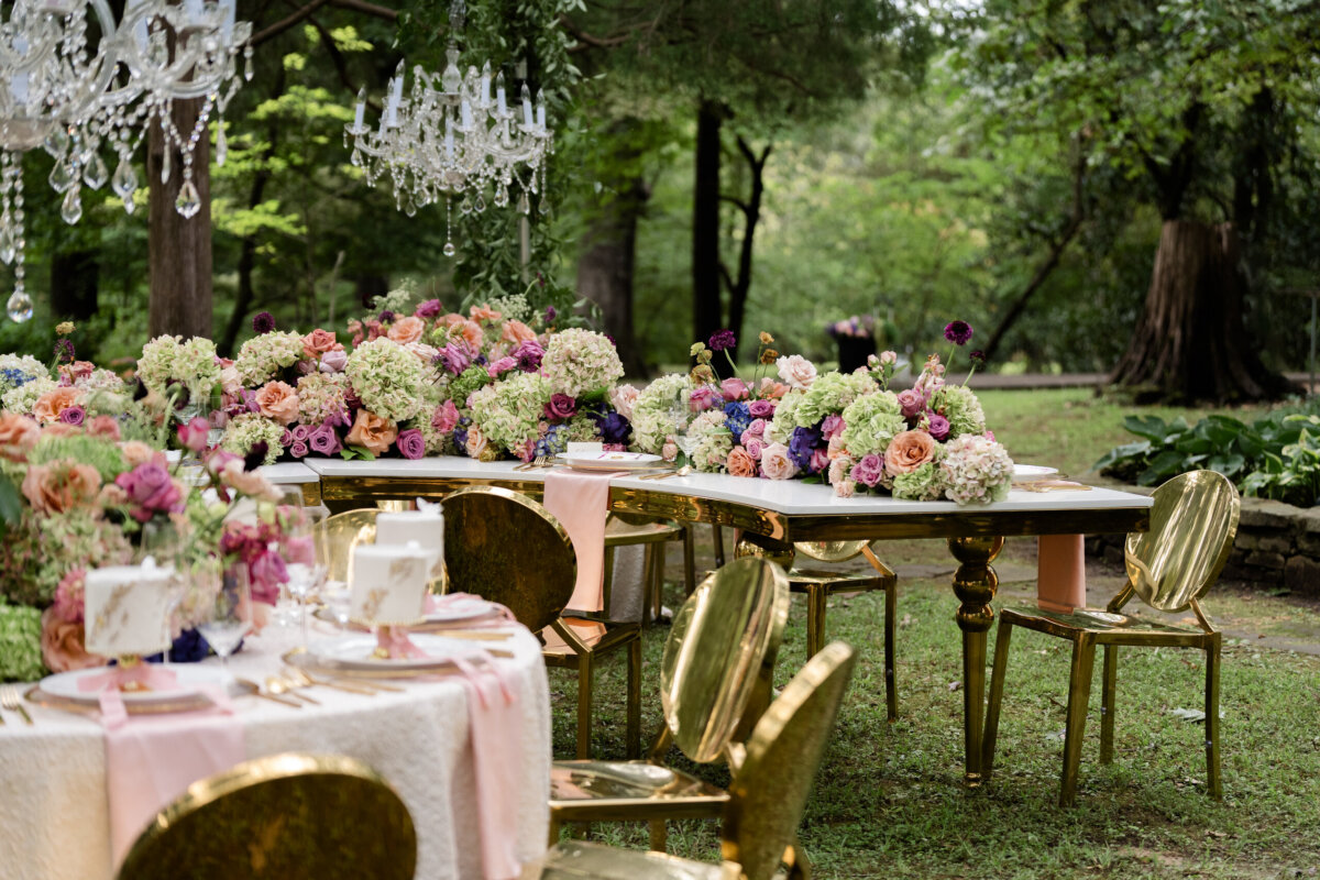The Top Event Spaces in Memphis - 2 Outdoor banquet tables decorated with large floral centerpieces and crystal chandeliers, set with gold chairs on a grassy lawn at one of the premier event venues in Memphis.