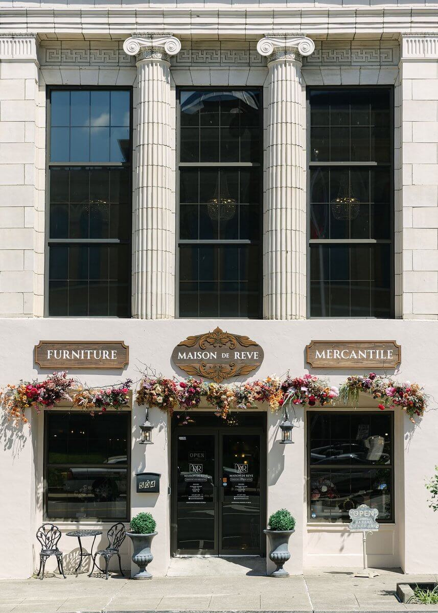 The storefront of Maison de Reve, a home decor store, with floral arrangements decorating the exterior and large windows framed by stone columns.