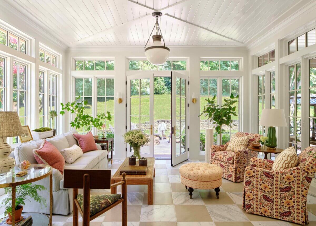 Bright sunroom with large windows, white ceiling, patterned armchairs, a sofa with pillows, potted plants, a round ottoman, and a central pendant light; expertly styled by some of the Best Interior Designers in Nashville. Doors open to a patio and green lawn.