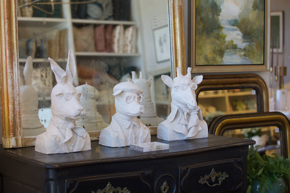Three white sculptural animal busts—a rabbit, a dog, and a giraffe—wearing glasses and scarves, displayed on a wooden dresser found at the best furniture and antique stores in Nashville.