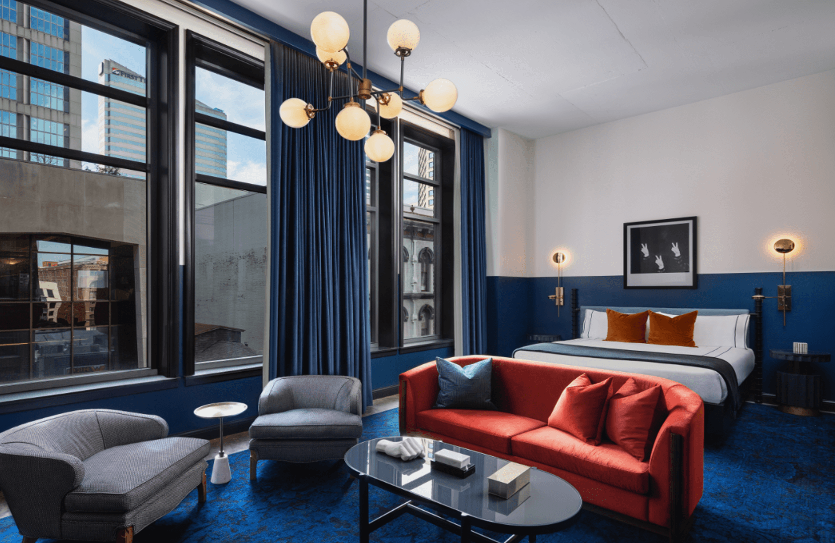 Top Luxury Hotels in Nashville - 18 Modern hotel room with large windows, a king bed, red sofa, gray chairs, blue carpet, round coffee table, and contemporary lighting. Enjoy city views outside—experience one of The Best Hotels in Nashville.