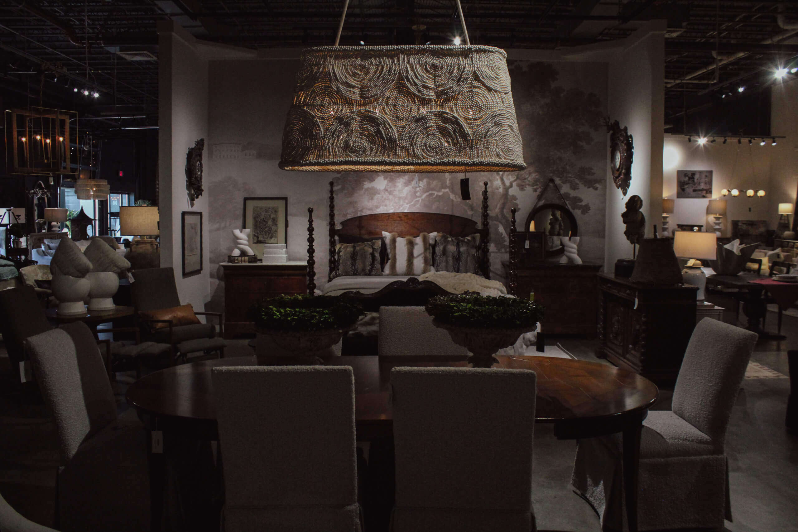 The Top Interior Designers in Memphis - 2 A dimly lit furniture showroom, inspired by the best interior designers in Memphis, features a large bed, various chairs, tables, and a prominent woven chandelier in the foreground.