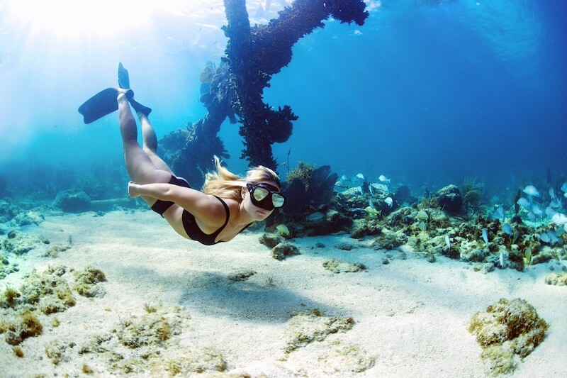 8 Adventurous Girls' Trips (All Skill Levels!) - 11 An adventurous woman wearing swim goggles and fins swims underwater near a sunken structure, surrounded by fish and coral—a perfect scene for unforgettable Adventurous Girls Trips.