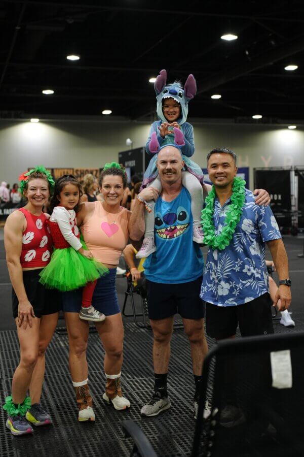 Meet the Co-Founder of Birmingham's "Recovery House" - 5 A group of five adults and two children, including Kelly Nguyen, in colorful costumes pose and smile together indoors at a gym or event space for an RCVRY House event.