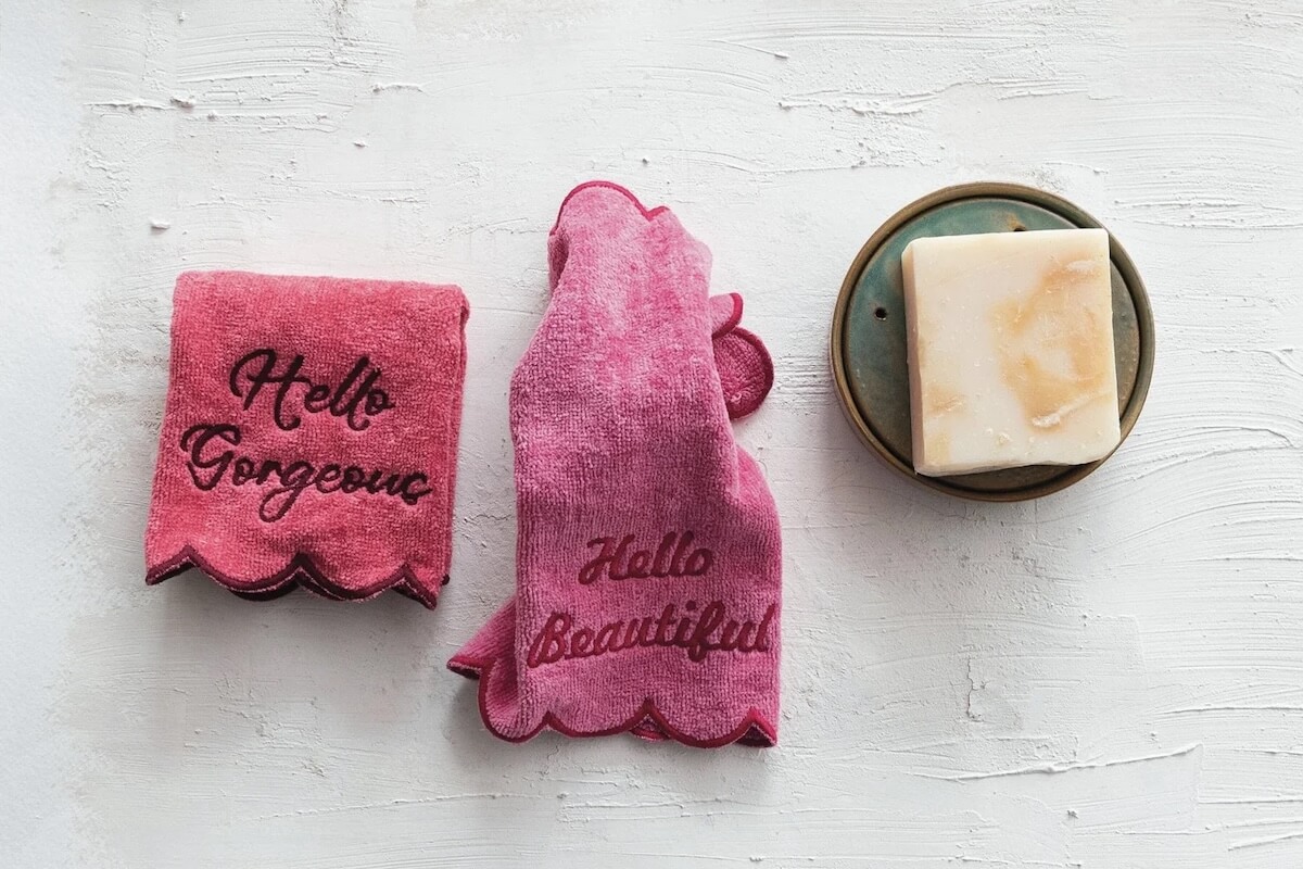 Two pink washcloths with scalloped edges and embroidered phrases "Hello Gorgeous" and "Hello Beautiful" sit next to a soap bar on a round dish, creating the perfect Winter Reset vibe for your Nashville home on a crisp white surface.